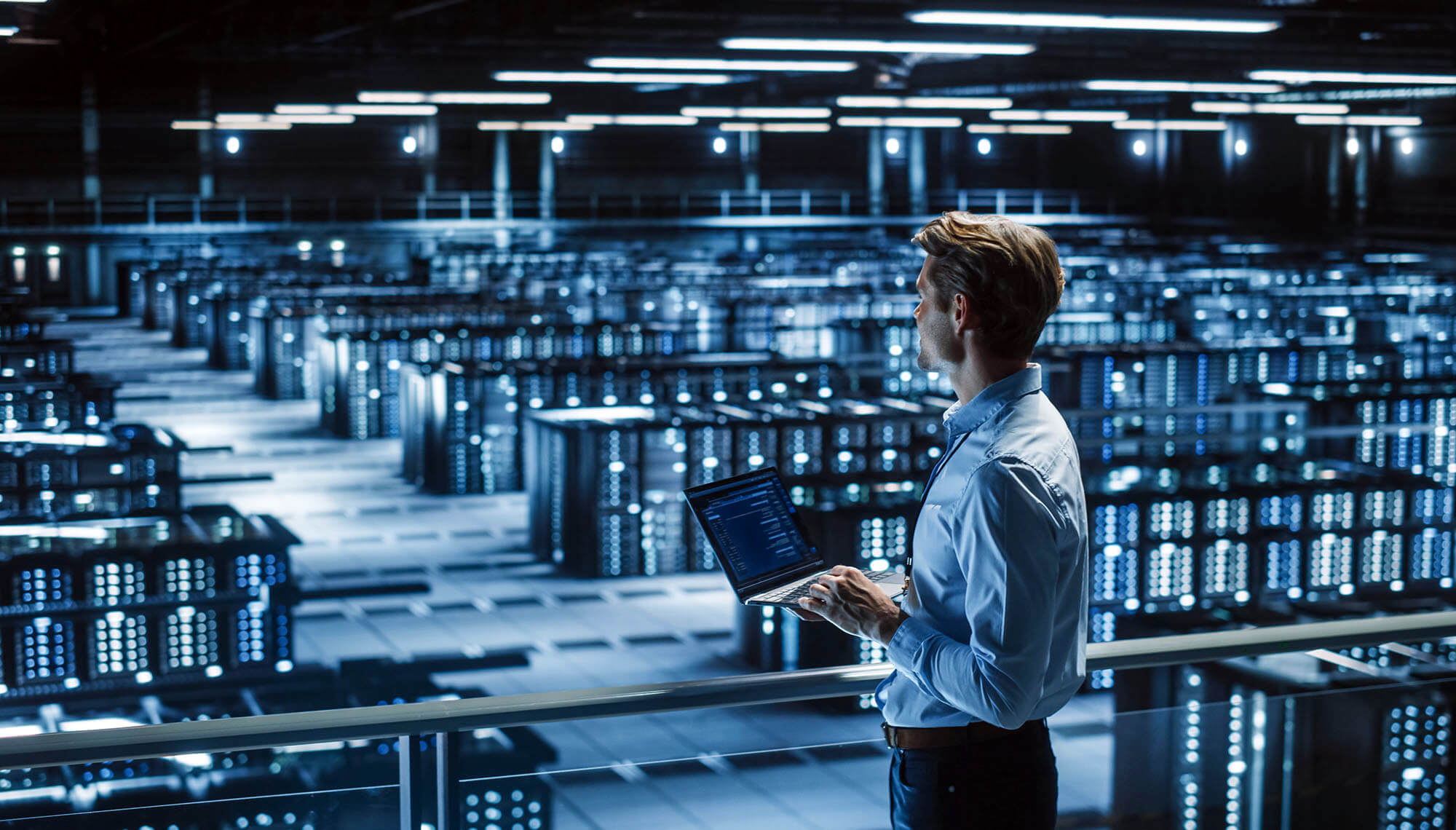 Osoba s notebookem hledící na řady serverových racků ve velkém, jasně osvětleném datovém centru.